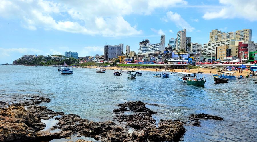 Orla do Rio Vermelho com barcos ancorados e prédios de Salvador ao fundo.